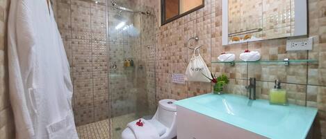 A bathroom with a glass shower enclosure, a toilet with a white towel, a sink with a glass countertop, and a mirror above the sink.