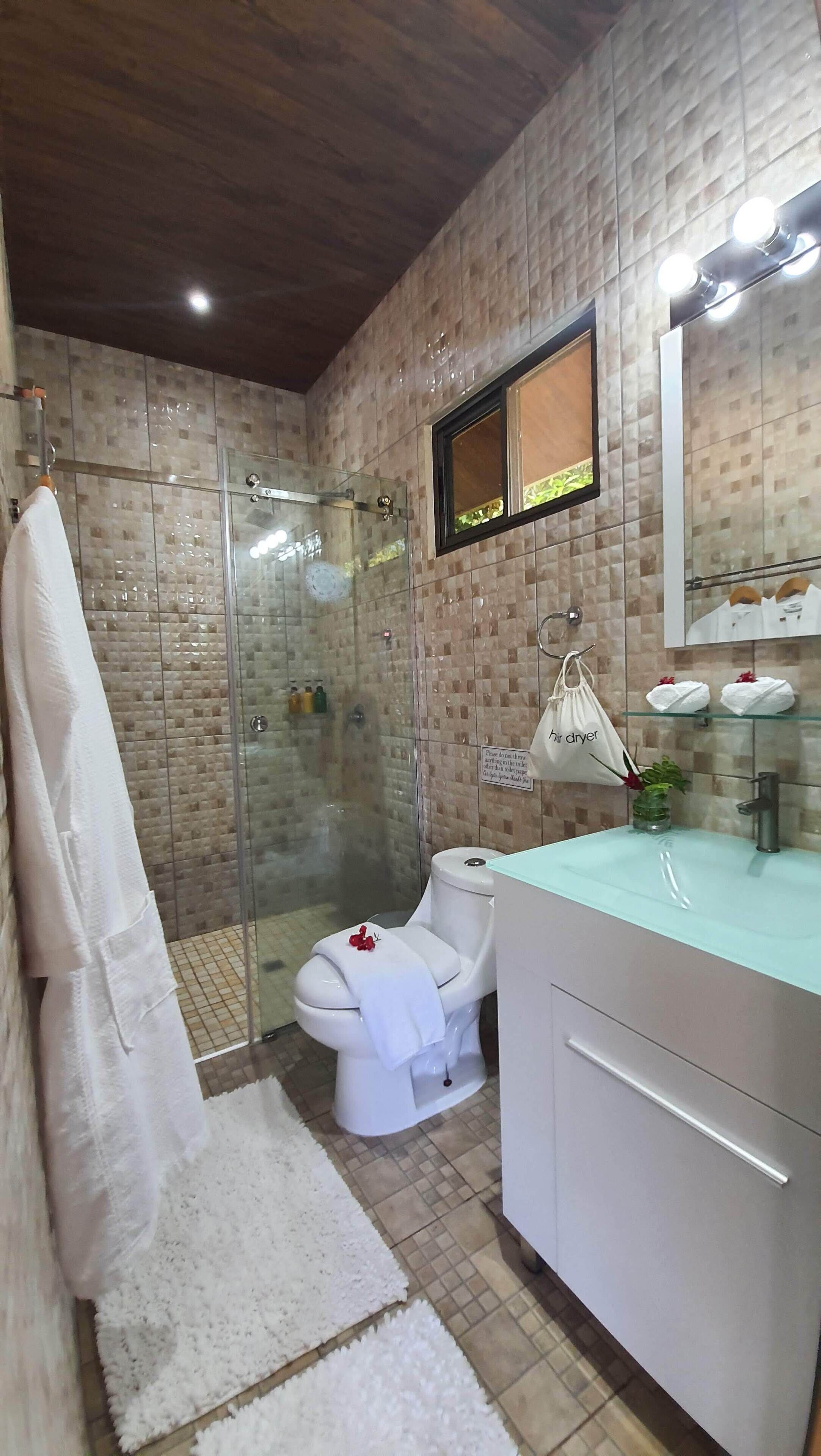 Un baño con ducha envuelta en vidrio, inodoro con toalla blanca, lavabo con repisa de cristal y espejo sobre el lavabo.