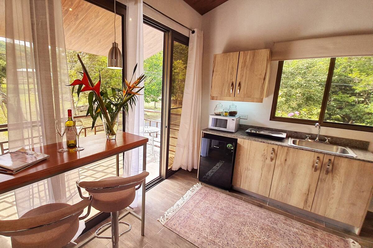 A modern kitchen with wooden cabinets, a microwave, and a sink. There is a dining area with two chairs and a table, and a window offering a view of greenery.