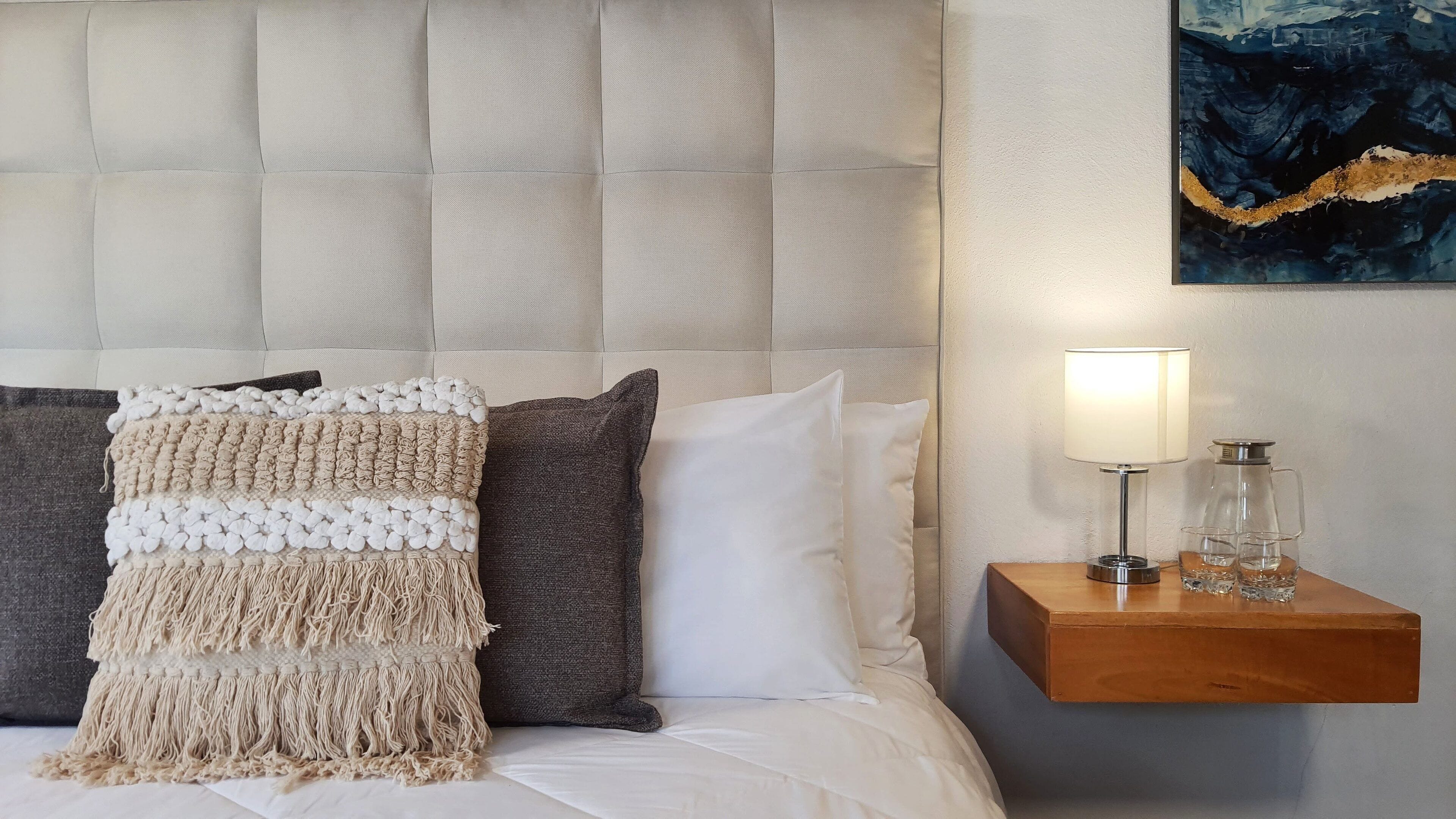 Una cama impecablemente tendida con una manta de macramé, una almohada gris y una blanca. Junto a la cama, una mesita de noche de madera con una lámpara y dos vasos.