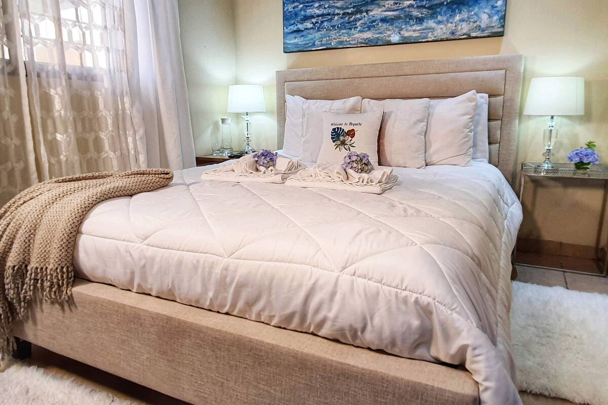 A neatly made bed with a beige quilt, two pillows with a floral design, and a welcome sign.
