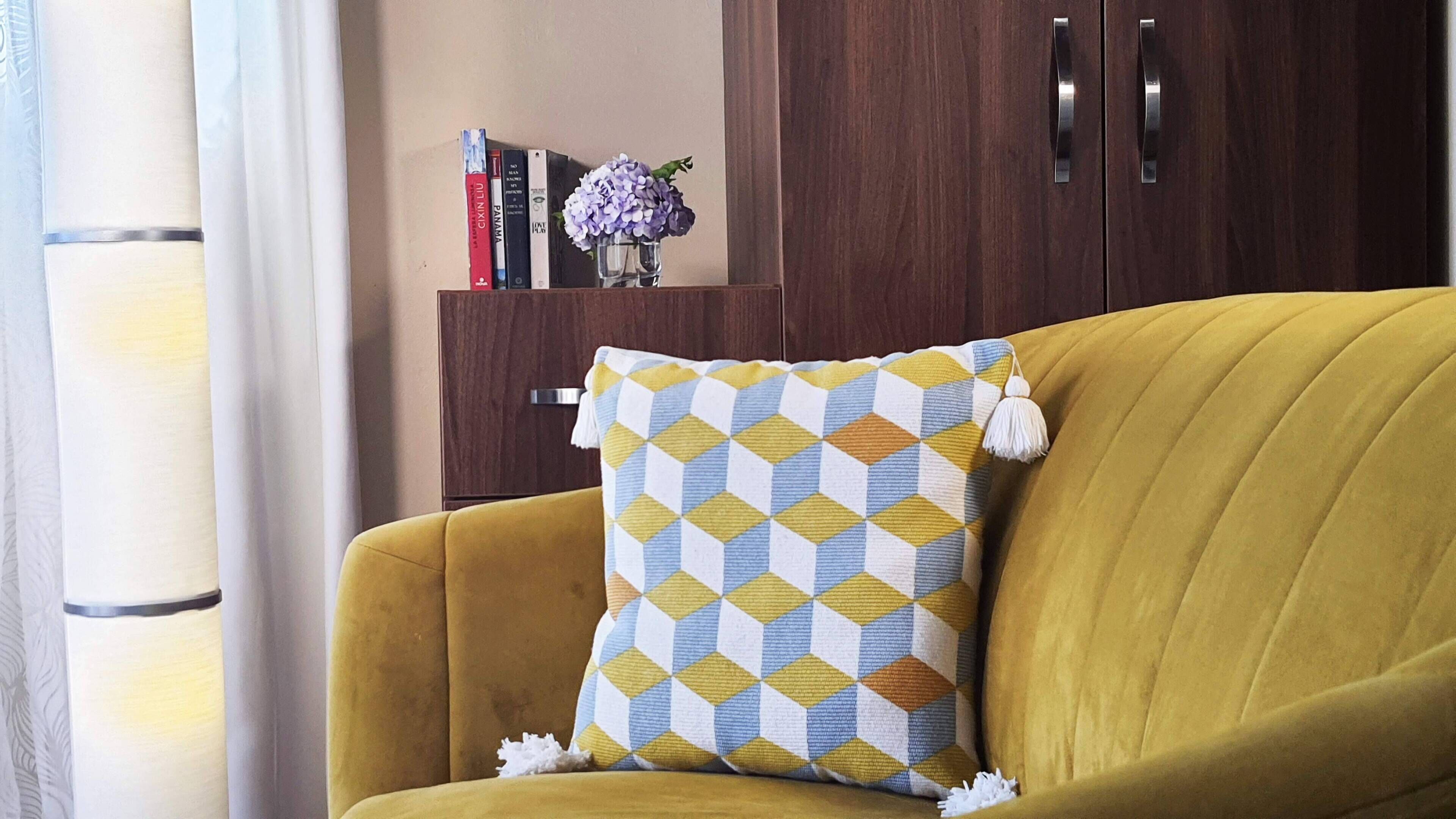 A mustard-colored armchair with a patterned cushion and a small vase with purple flowers on a wooden cabinet.