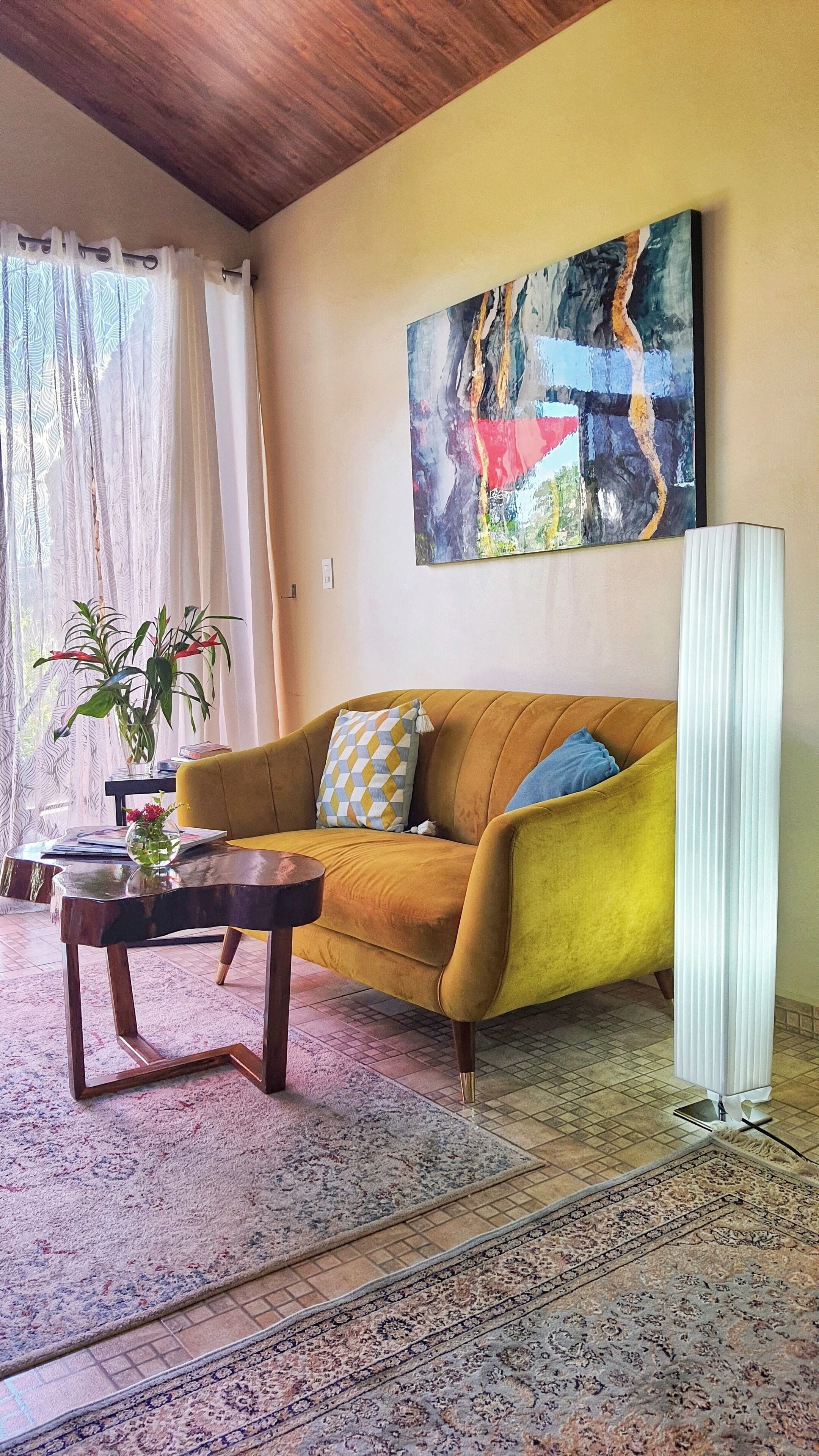 A living room with a mustard sofa, a coffee table, a floor lamp, a painting, and a potted plant.