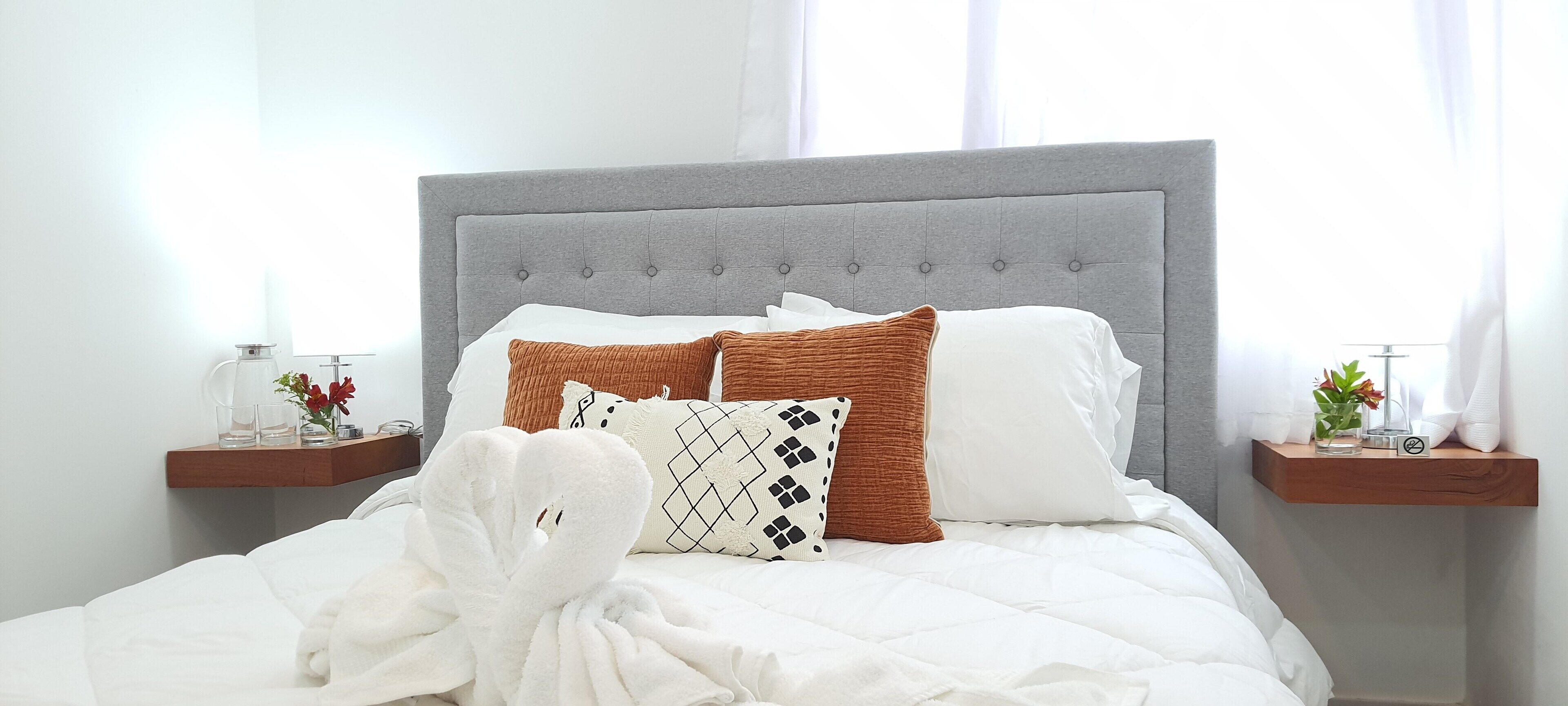 A neatly made bed with a grey upholstered headboard, white bedding, and a variety of patterned pillows. There are two bedside tables with decorative items and a vase of flowers.