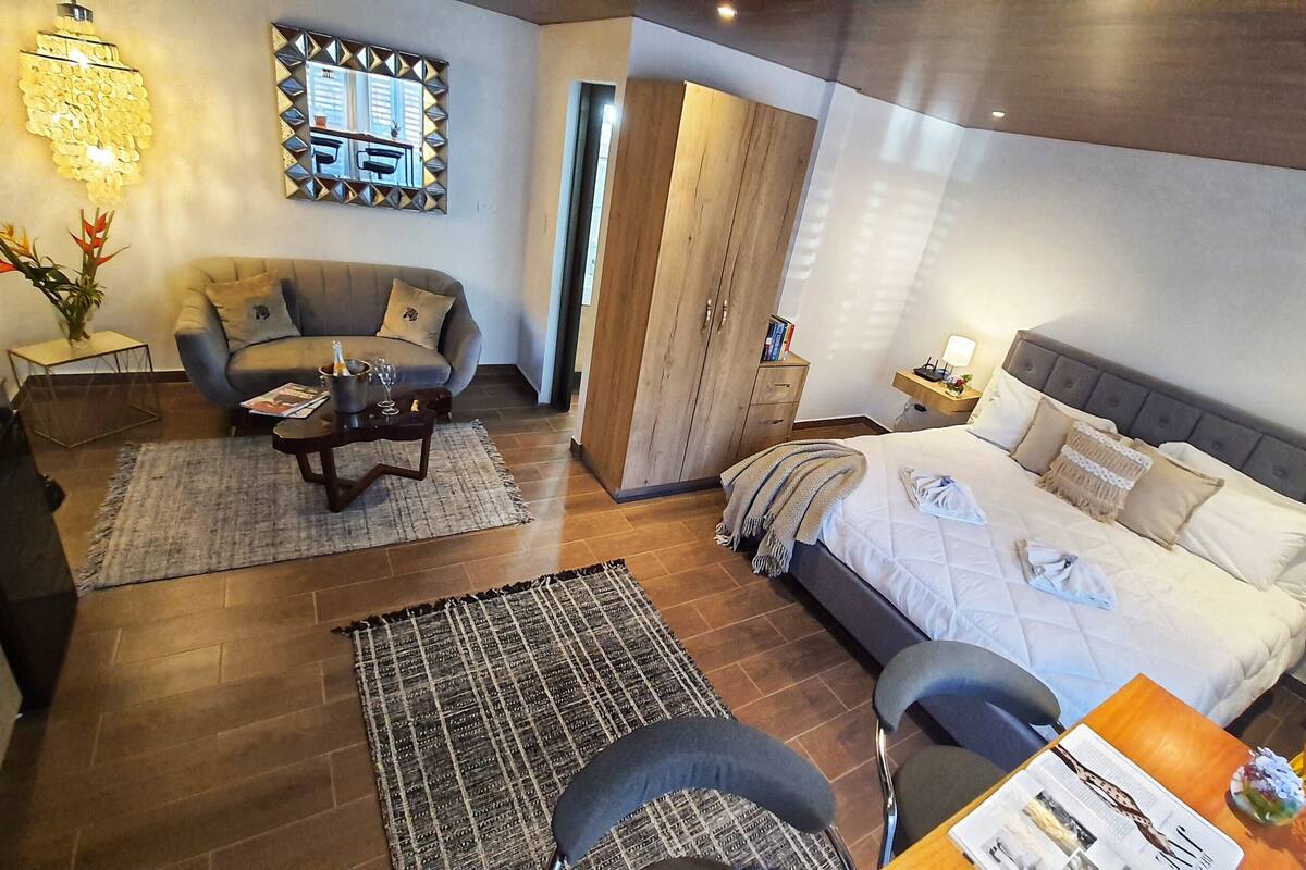 A modern hotel room with a bed, a sofa, a small table, and a chair. There is a mirror on the wall and a wooden wardrobe.