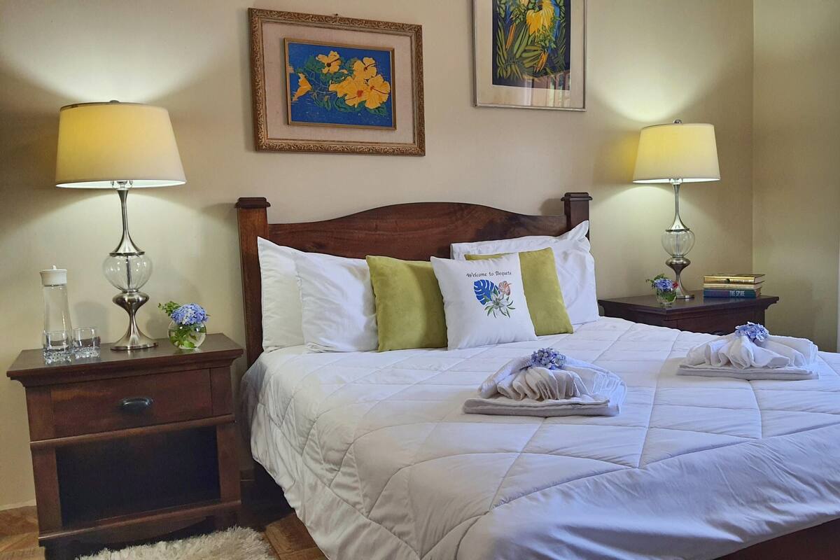 A neatly made bed with white linens, two bedside tables with lamps, a framed floral painting, and a window with sheer curtains.