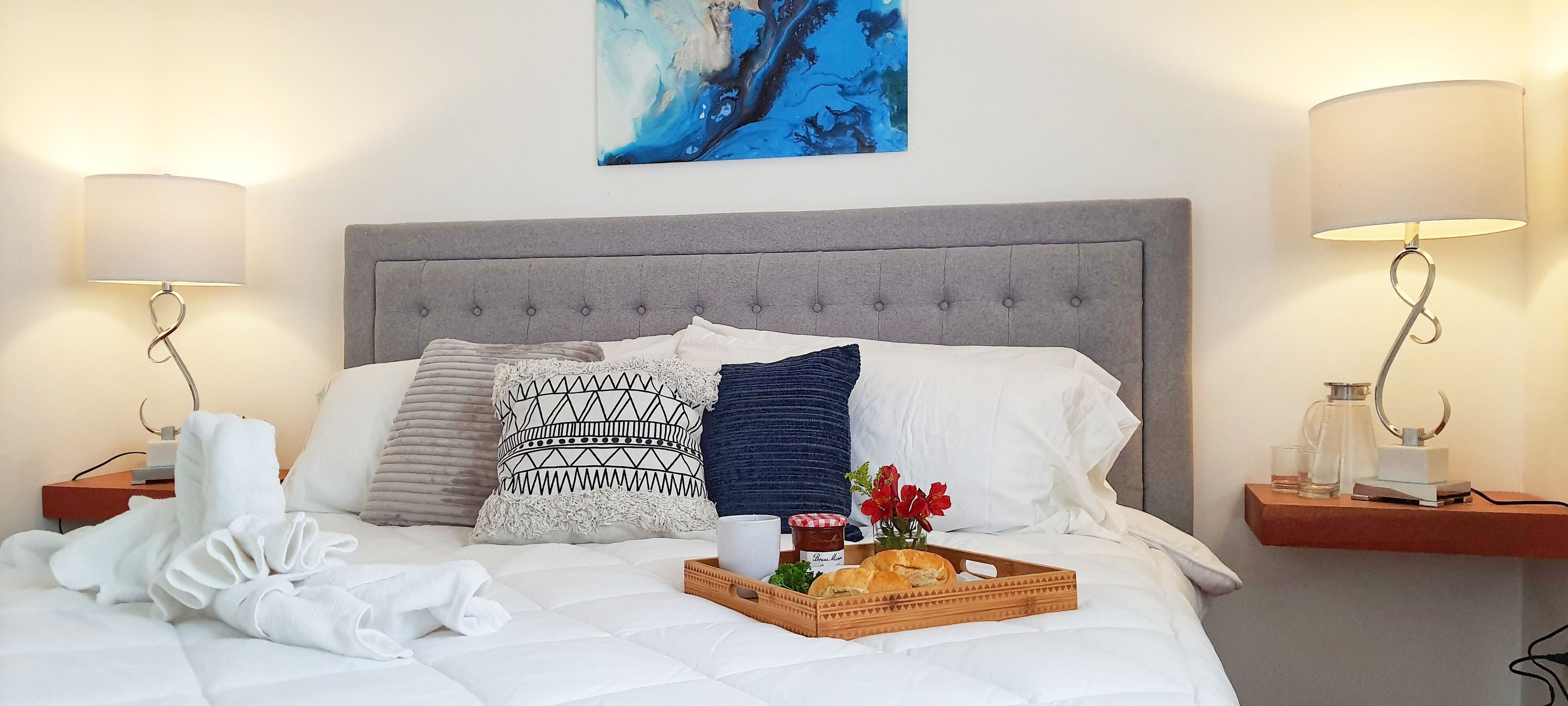 Un lit bien fait, avec une tête de lit grise, deux tables de chevet avec des lampes, un plateau avec du pain et des fleurs, et un tableau accroché au mur.