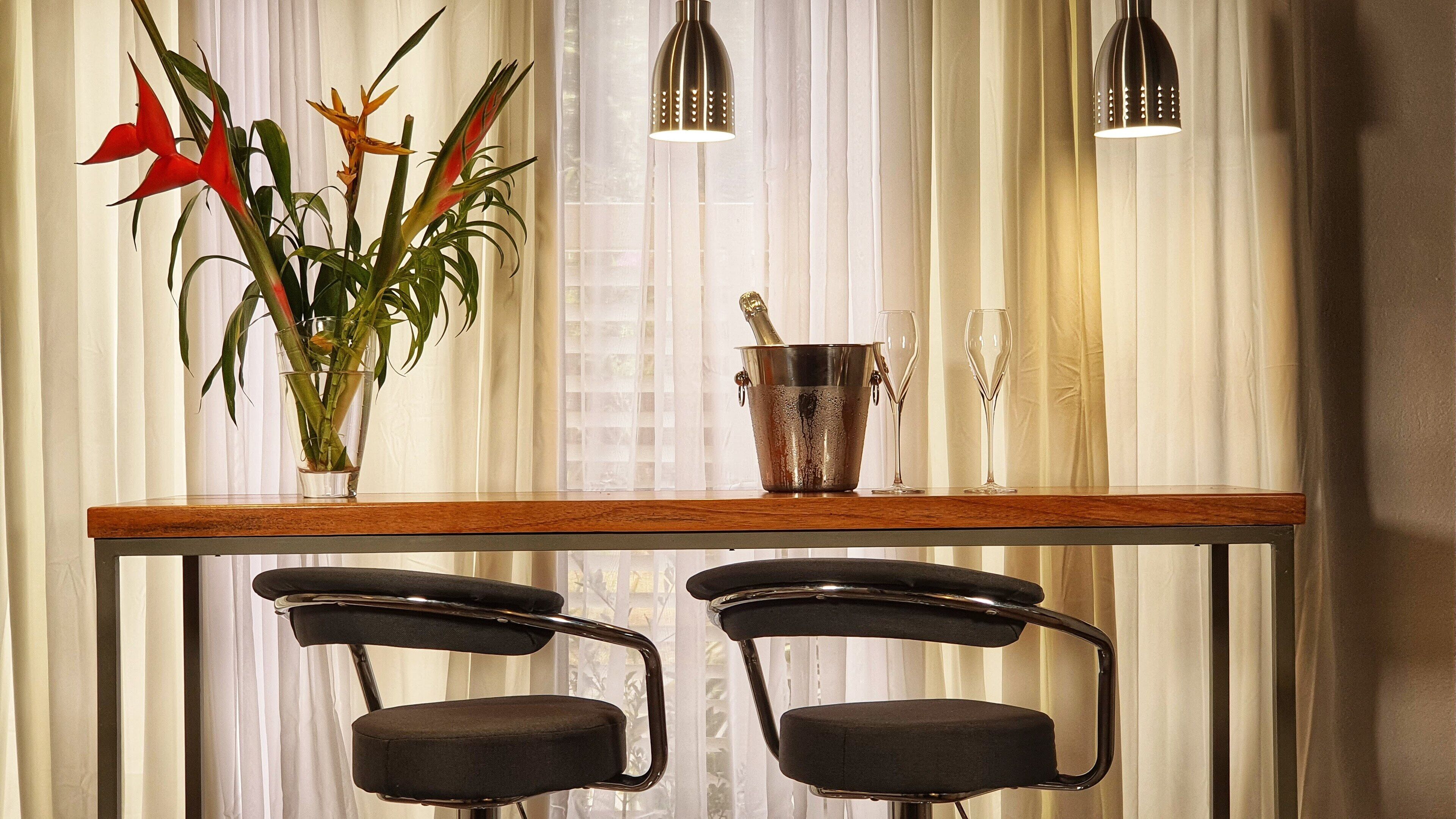A dining area with a wooden table, two black chairs, a vase with red flowers, a champagne bucket, and two wine glasses.