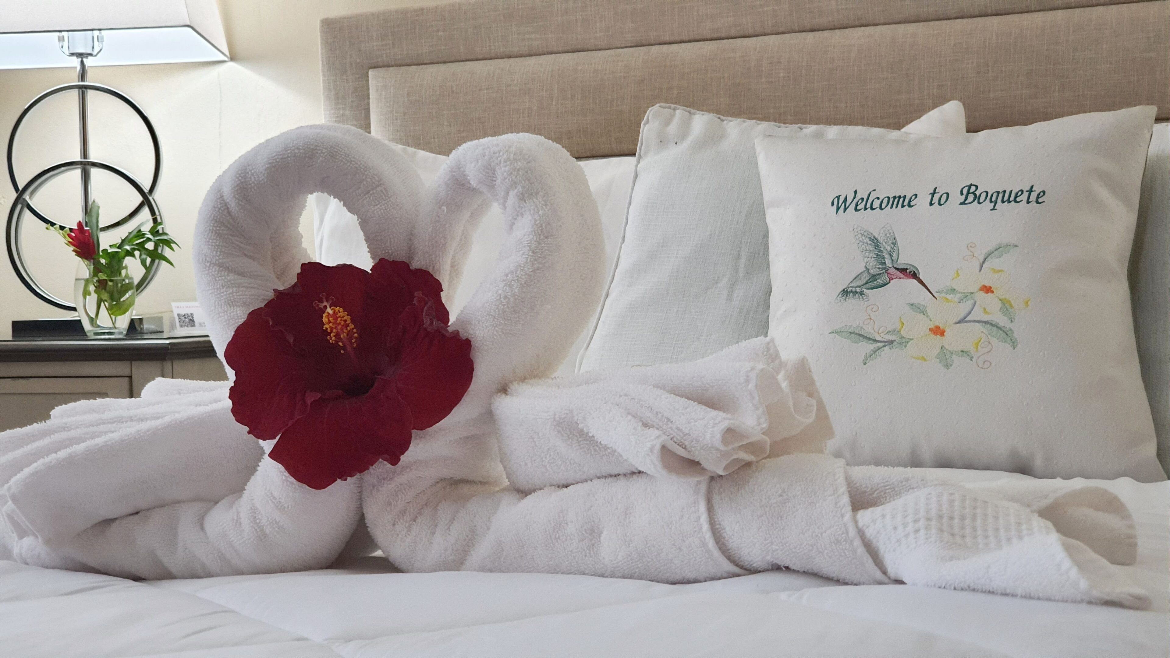 A neatly made bed with white towels arranged in a heart shape, a red hibiscus flower, and a pillow with a bird and floral design.