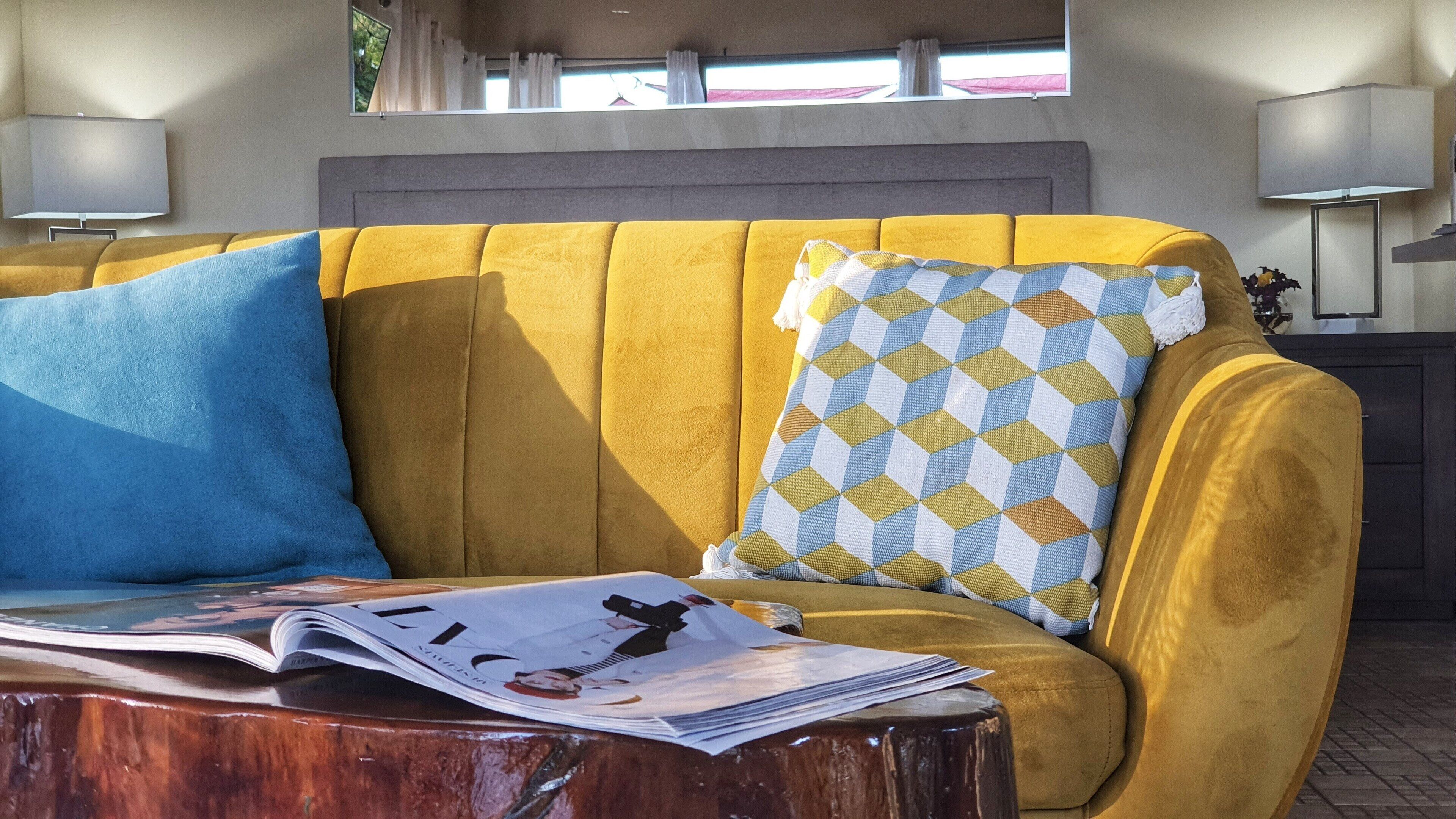 A yellow sofa with a blue and yellow patterned pillow, a wooden coffee table with a magazine, and a window with curtains.