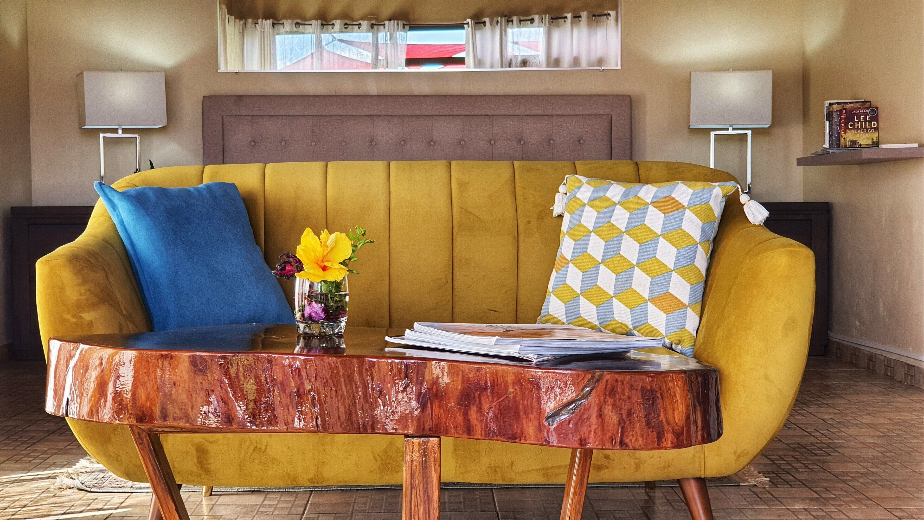 Un sofá amarillo con almohadones azules y estampados, una mesa de centro de madera con revistas, y una ventana con cortinas.
