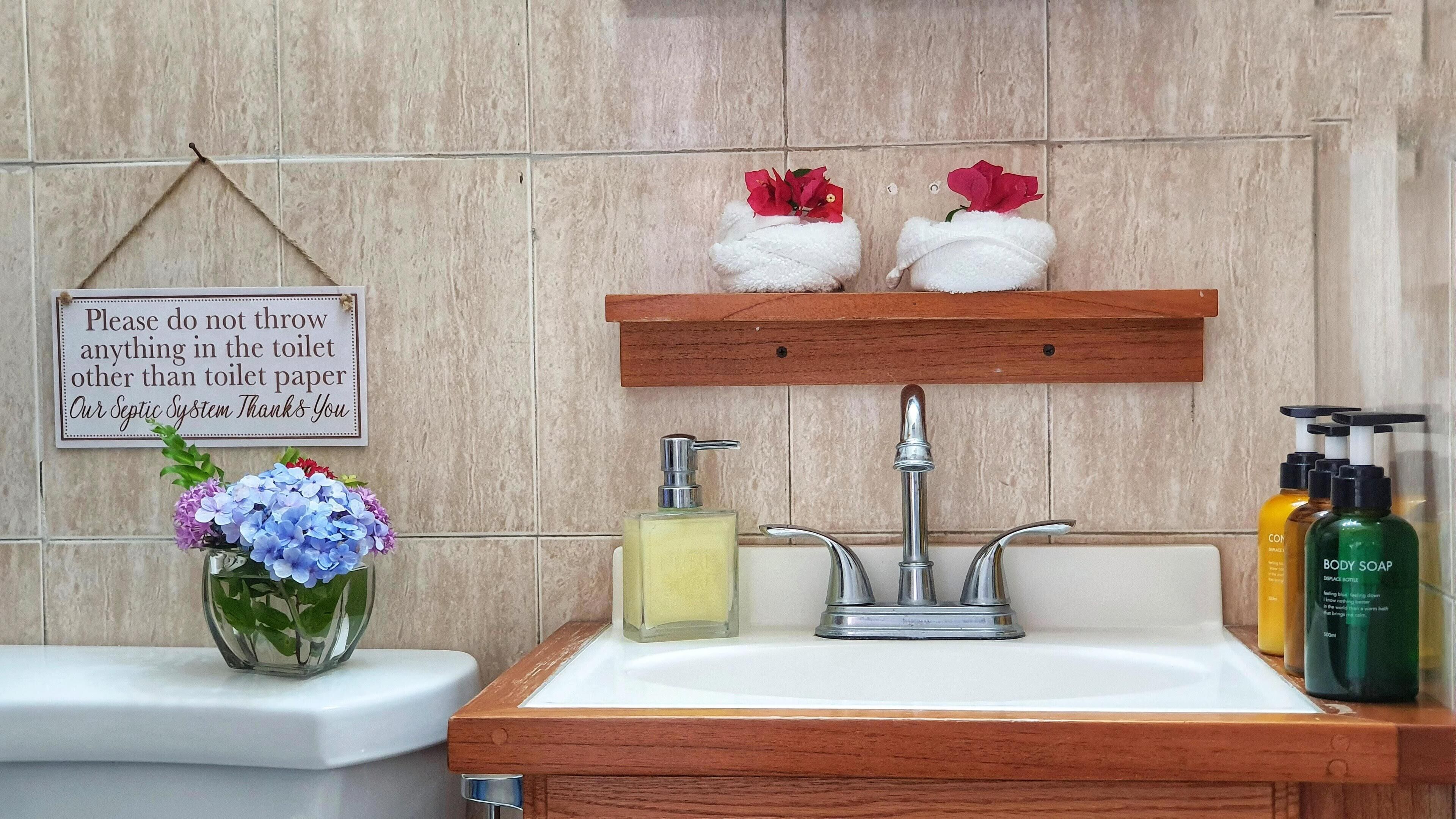 A bathroom with a toilet, a sink, and a shelf with toiletries. There is a sign on the wall and a vase with flowers on the sink.