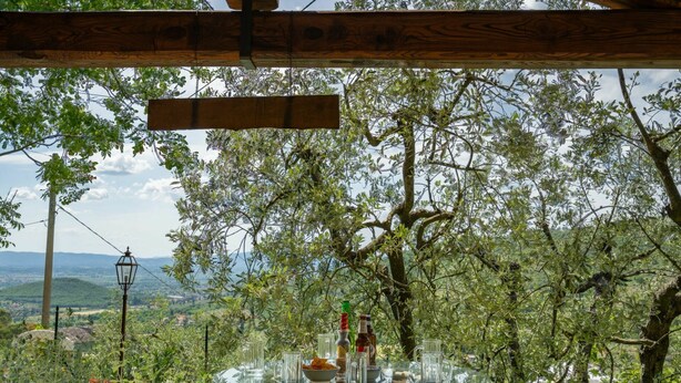 Un tavolo con piano in vetro, adornato con bottiglie e bicchieri, circondato da sedie di legno, con sullo sfondo alberi e un paesaggio lontano.