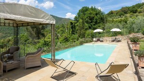 Area piscina con lettini, zona relax coperta e vista su un paesaggio montuoso.