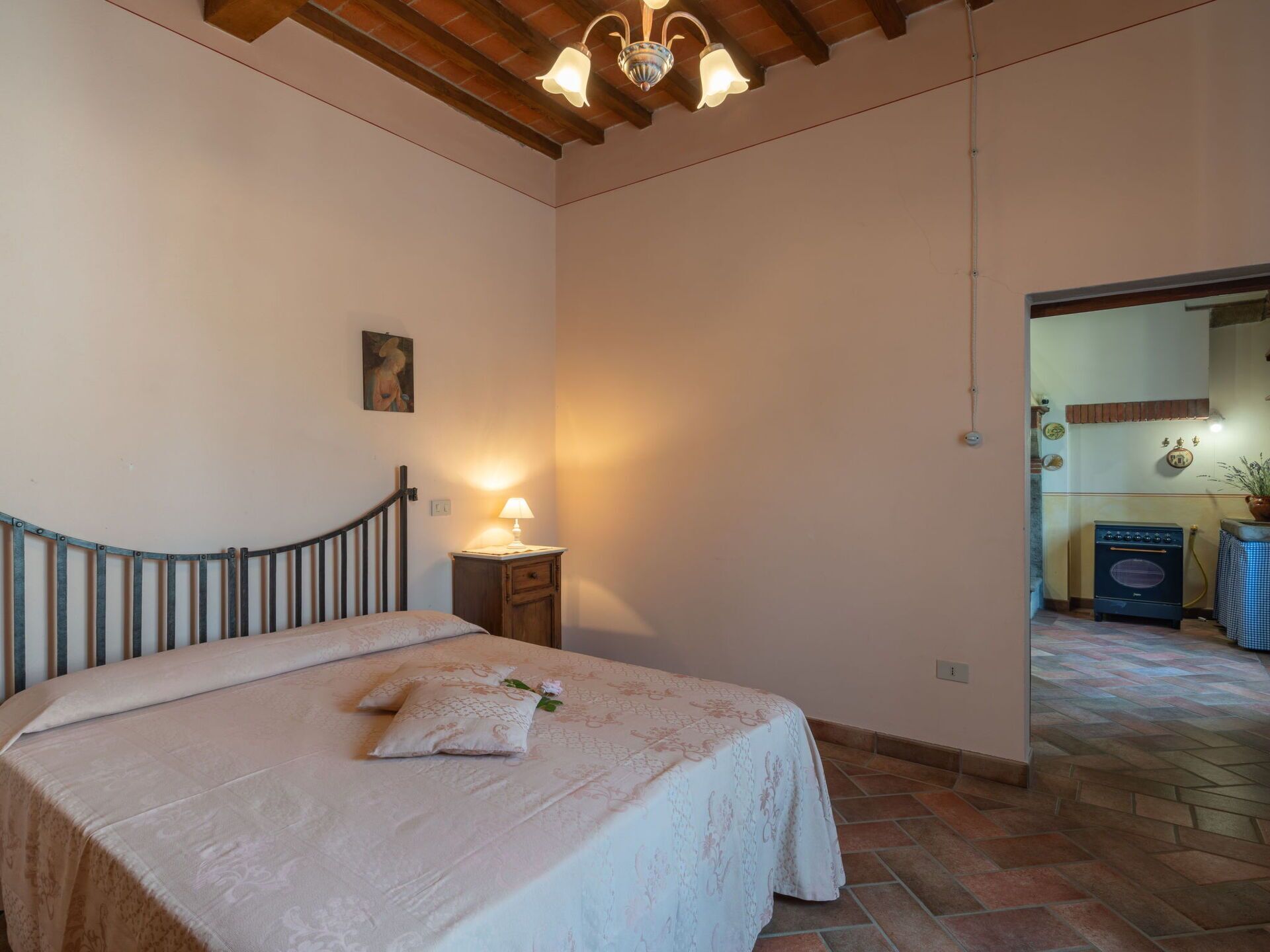 A bedroom with a bed, a nightstand with a lamp, a wooden ceiling, and a view into another room.