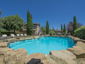 Una piscina azul clara rodeada de rocas, con sillas de descanso y un edificio de piedra al fondo.