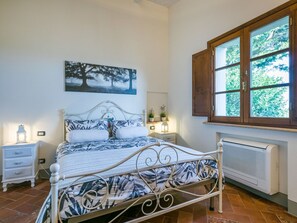 Un dormitorio con cama, mesitas de noche, un radiador, una ventana con contraventanas de madera y un cuadro en la pared.