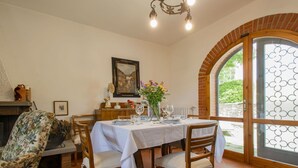 Un coin repas avec une table dressée pour quatre personnes, des chaises, une cheminée et une grande fenêtre donnant sur un paysage verdoyant.