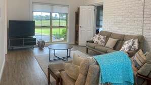 A living room with a TV, sofa, and a view of a green landscape through a large window. - The 19th Green - Located near the golf course! Pet Friendly - Outside Only (Robe)