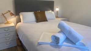 A neatly made bed with white linens, a brown pillow, and a rolled white towel on the bed. There are two bedside tables with lamps and a drawer unit with three drawers. - The 19th Green - Located near the golf course! Pet Friendly - Outside Only (Robe)