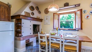 A kitchen with a white refrigerator, wooden cabinets, a dining table with chairs, patterned floor tiles, and a window with a view of greenery.