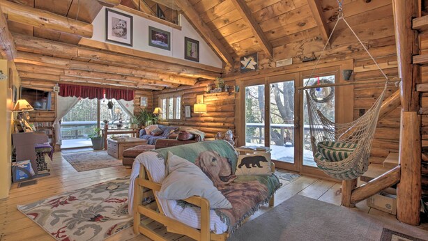 Interior acogedor de una cabaña de troncos con una cama de madera, una hamaca y un amplio ventanal con vistas a un paisaje nevado.