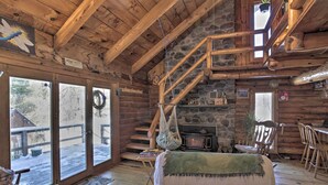 Interior acogedor de una cabaña con chimenea de piedra, escalera de madera y un gran ventanal con vista a una terraza nevada.