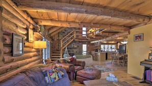 Interior de una cabaña de troncos con techo de madera, una escalera de piedra, una sala de estar con mobiliario de cuero y una zona de cocina.