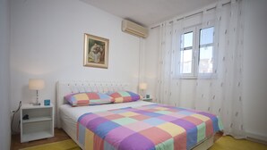 Une chambre à coucher comprenant un lit, deux tables de chevet, une commode, un climatiseur fixé au mur, une fenêtre avec des rideaux et un tableau accroché au mur.