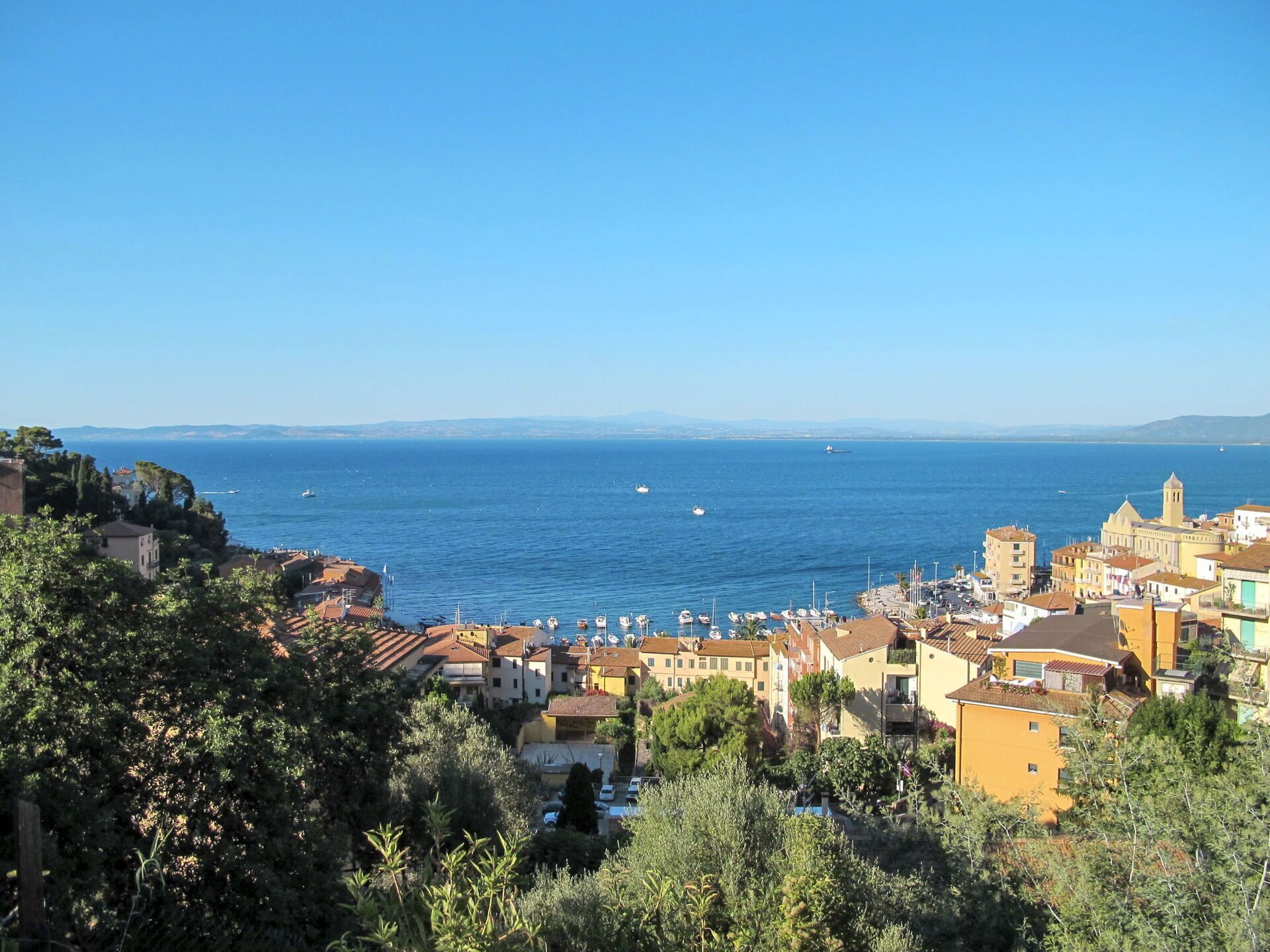 A coastal town with buildings along the sea, boats docked, and a clear blue sky.