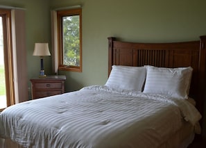 Un dormitorio con cabecera de madera, dos almohadas blancas, una cama con manta a rayas, una mesita de noche con lámpara y una ventana con cortinas.