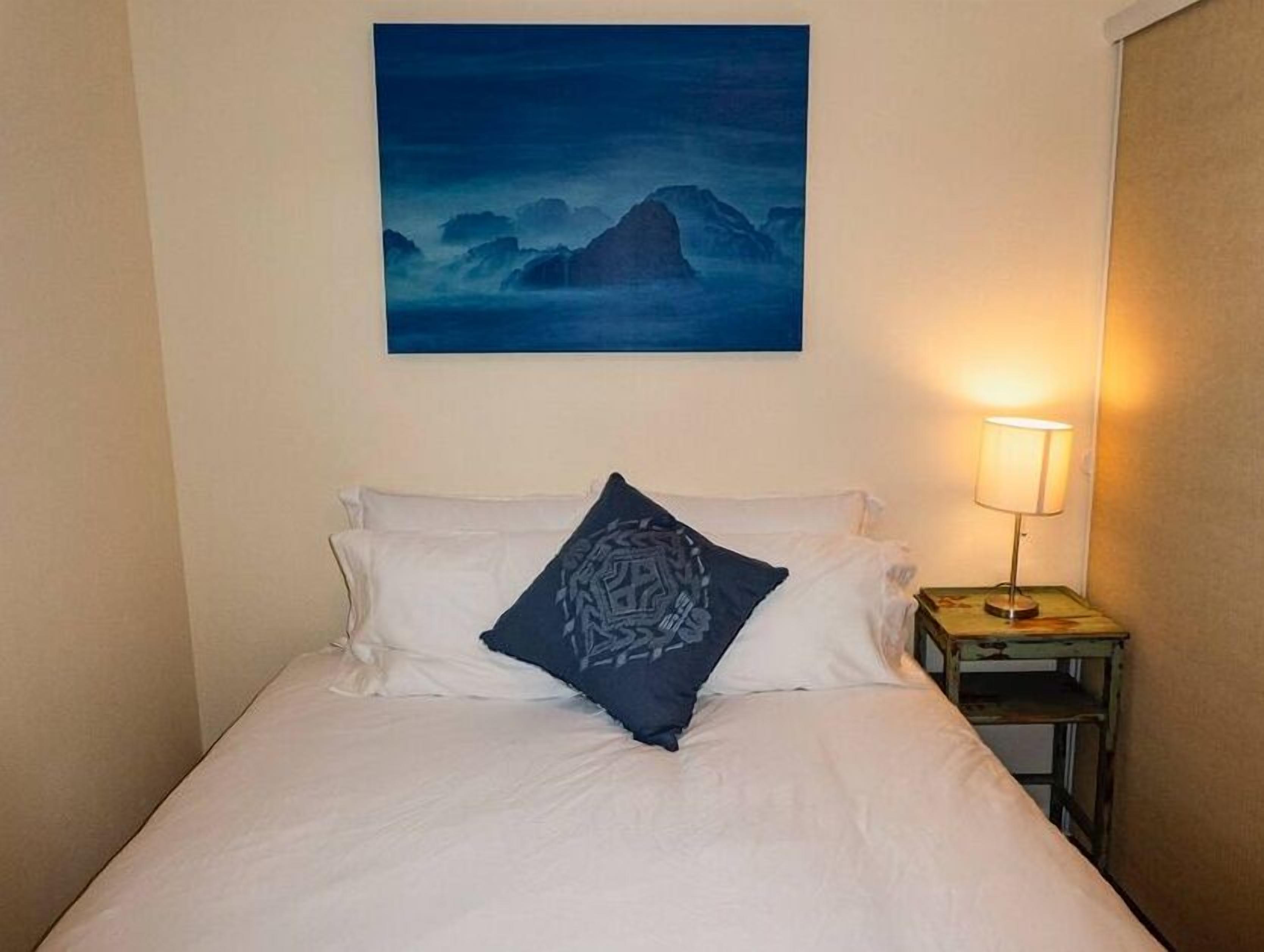 Un lit bien fait, avec une literie blanche et un seul coussin décoratif, une table de chevet avec une lampe et un tableau représentant un paysage de montagne accroché au mur.