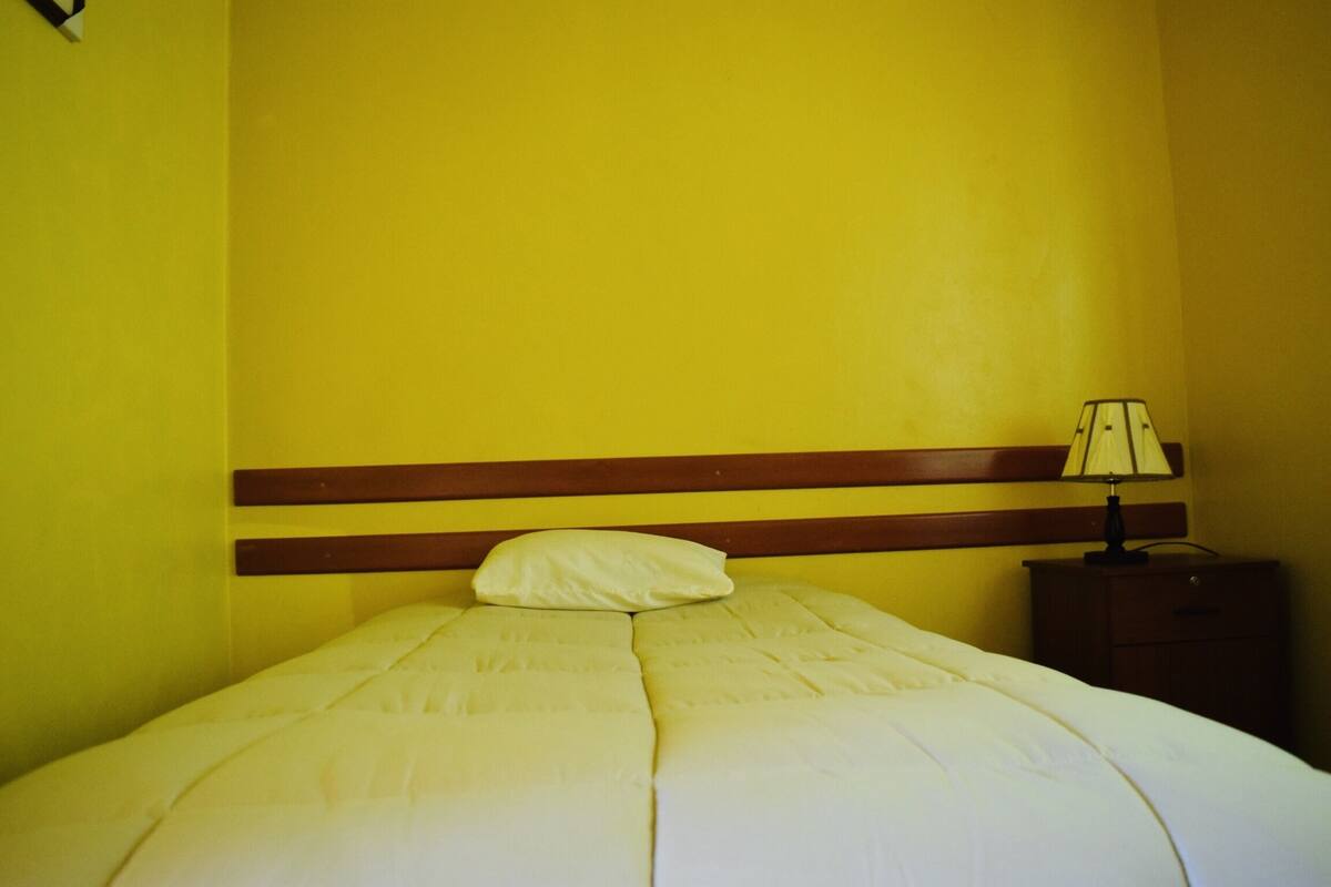 Una cama con cabecera de madera, almohada blanca y mesita de noche con lámpara.
