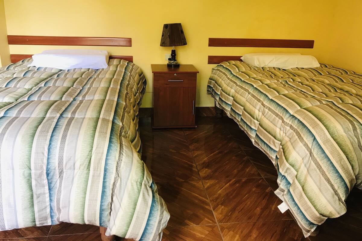 Habitación de hotel con dos camas, cabeceros de madera, una mesita de noche con lámpara y suelo de madera.
