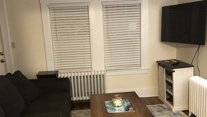 Una sala de estar con un sofá de color oscuro, una mesa de centro de madera con objetos decorativos, una alfombra estampada, un radiador y un televisor montado en la pared.