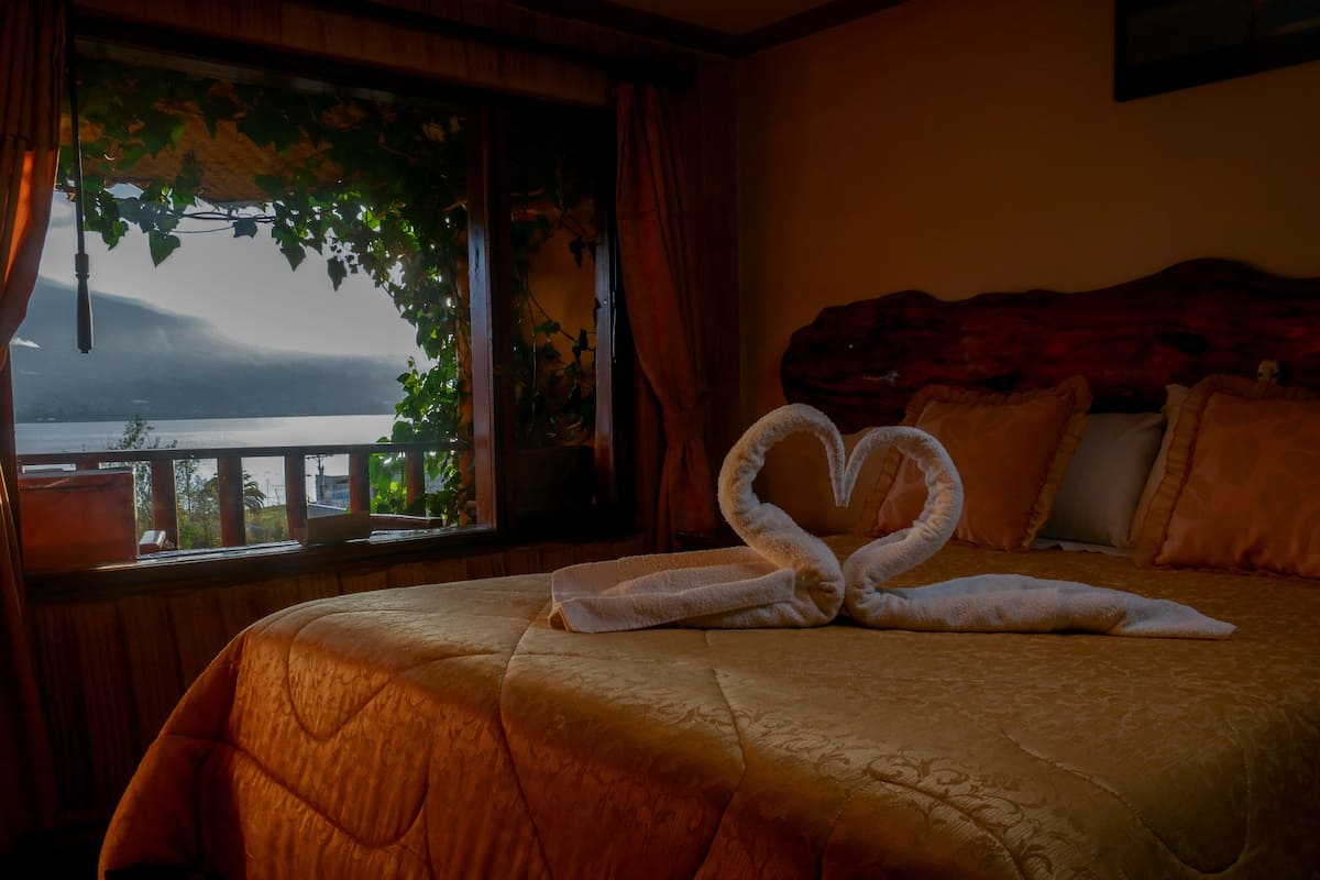 Habitación de hotel con una cama, una toalla dispuesta en forma de corazón, vistas a un cuerpo de agua y un balcón con plantas.