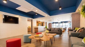 A modern lounge area with a flat-screen TV mounted on a white brick wall, wooden round tables, and comfortable seating including a grey sofa and red ottoman. - The Home Hotel (Bangkok)