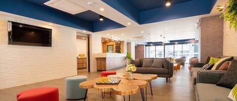 A modern lounge area with a flat-screen TV mounted on a white brick wall, wooden round tables, and comfortable seating including a grey sofa and red ottoman.