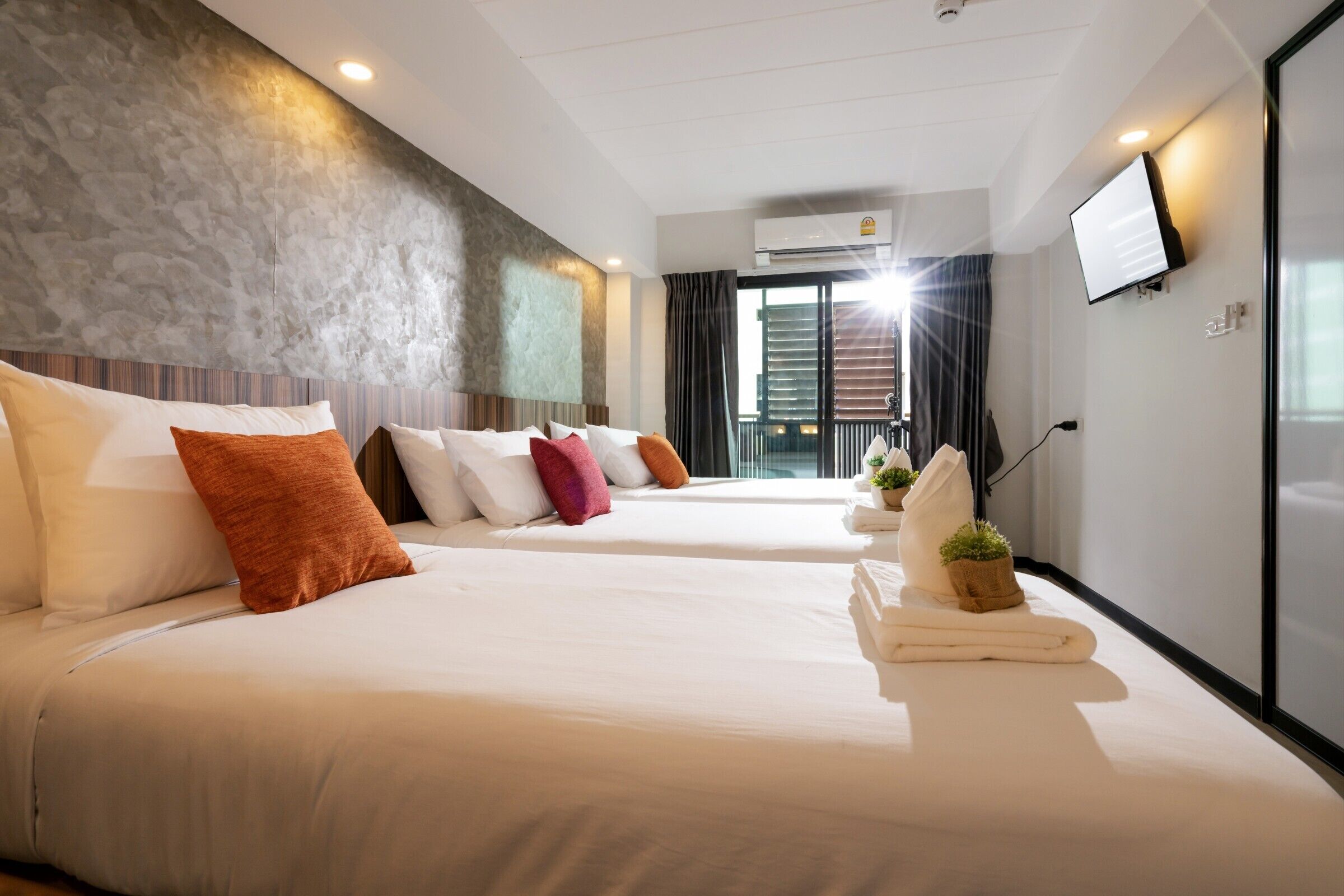 A hotel room with two beds, a flat-screen TV, an air conditioning unit, and a balcony with curtains.