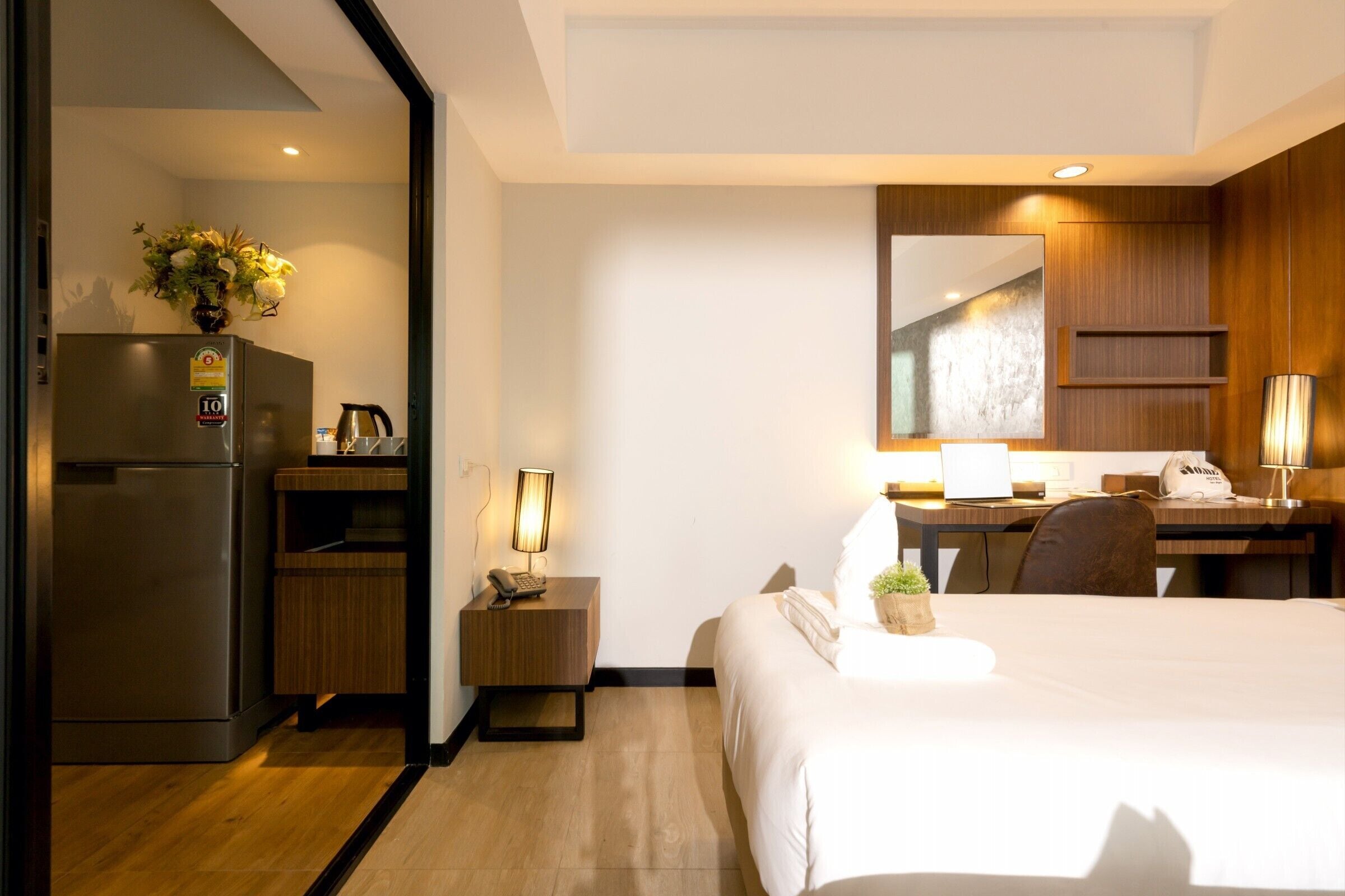 A hotel room with a bed, a desk with a chair, a refrigerator, a bedside table with a lamp, and a mirror on the wall.