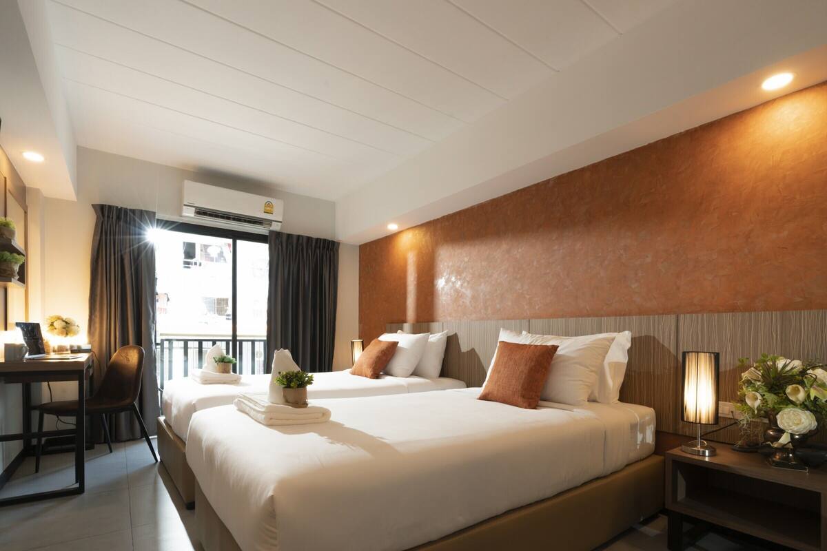 A hotel room with two beds, a desk, a chair, a nightstand with a lamp, and a view of the outside.