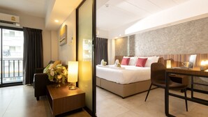 A hotel room with a bed, a desk with a laptop, a chair, a sofa, a nightstand with a lamp, and a view of the outside. - The Home Hotel (Bangkok)