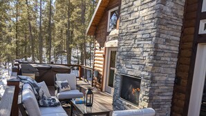 A wooden deck with outdoor furniture, a stone fireplace, and a view of a forested area. - Whispering Pines - LUXURY Cabin-Hot Tub, Fireplace, Huge Deck, Pool Tbl, 6 Acres (Florissant)