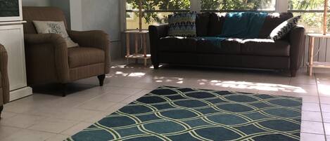 A living room with a sofa, armchair, and a patterned rug. There are windows with a view of trees outside.