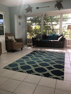 A living room with a sofa, armchair, and a patterned rug. There are windows with a view of trees outside.