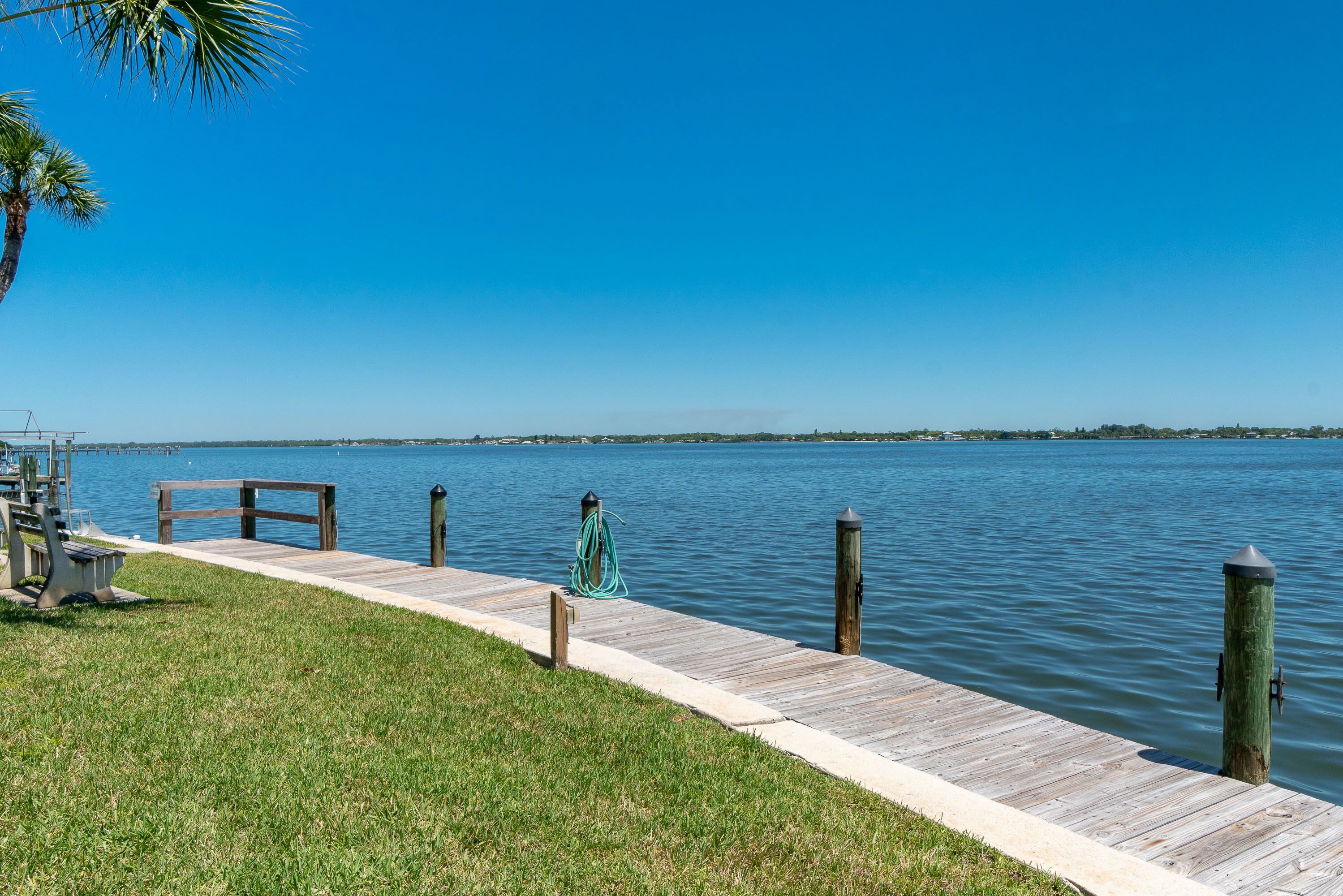 A wooden dock extending into calm water, with a grassy area and a palm tree on the left.