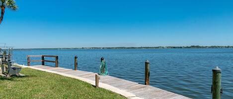 A wooden dock extending into calm water, with a grassy area and a palm tree on the left.