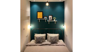 A bedroom with a teal accent wall, a bed with two patterned pillows, and wall-mounted shelves with framed pictures and a small plant. - Wonderloft Hostel Jogja (Yogyakarta)