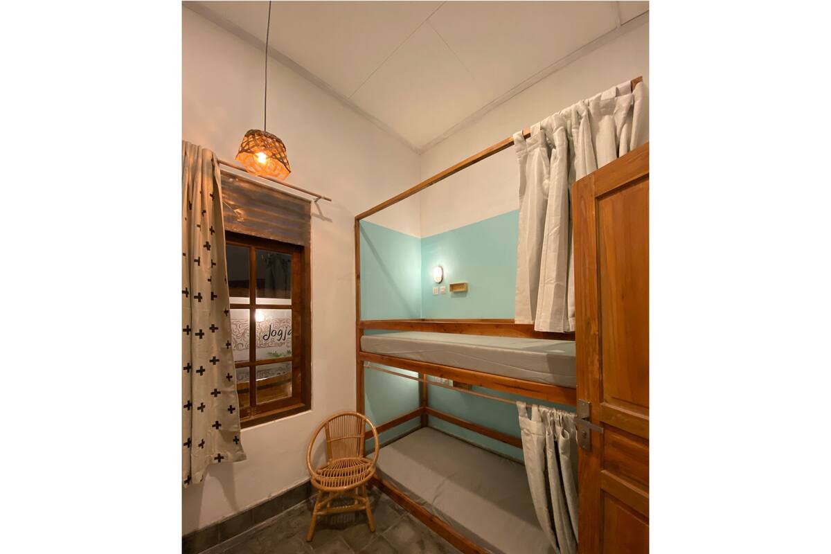 A small, cozy room with a bunk bed, a window with curtains, a wicker chair, and a wall-mounted light fixture.