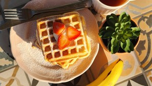 A plate with a waffle, a cup of tea, a fork, a banana, and a small potted plant. - Wonderloft Hostel Jogja (Yogyakarta)